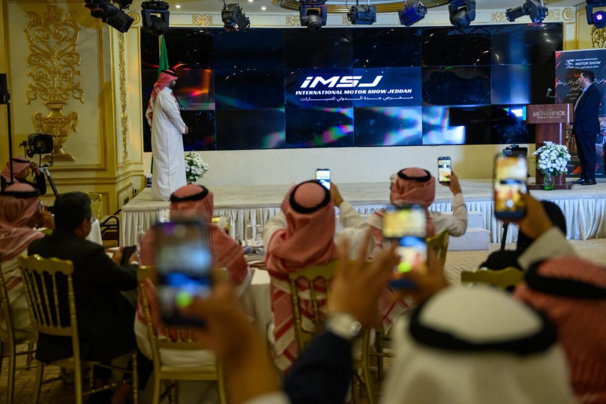 شعار IMSJ المعرض الدولي للسيارات بجدة على الشاشة خلال المؤتمر الصحفي والحضور يصورون بهواتفهم